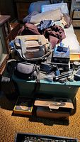 Full view of unorganized pile of camera bags, accessories, power cords, and boxes on green plastic storage bin and carpet floor