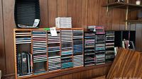 Three CD racks on a wooden surface against wood-paneled wall, filled with assorted CDs.