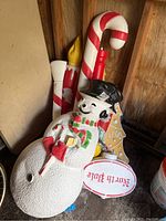 Photo showing large white plastic snowman lying down with a black hat and scarf, leaning on large candy cane decorations.