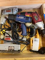 Multiple power tools including drills, handheld heater, sander, and soldering iron packed in wooden crate