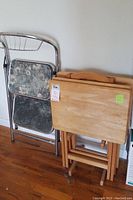 Side view of three wooden folding snack tables next to folding two-step metal ladder leaning against wall on hardwood floor.