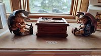 Main photo showing two Hummel figurines and the wooden music box in between, placed on a wooden surface near windows.