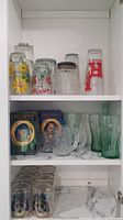 Shelf showing various glassware including floral and geometric print glasses, clear patterned mugs, Coca-Cola green tinted glasses, and Lord of the Rings glass goblets in boxes.