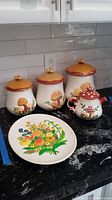 Photo showing three mushroom themed kitchen storage canisters and a matching mushroom themed teapot along with a floral decorative platter on a black countertop with gray tile backsplash.