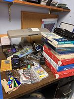 Wide view of desk surface showing a vintage desk lamp, multiple unopened reams of Avery and Highland printer paper stacked on the right, and assorted small office tools including label printer, tapes, and color swatches.