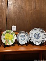 Three plates on wooden shelf with stands. Left plate: yellow apple design. Center plate: Blue Danube, scalloped edge, blue floral. Right plate: Blue Danube, pierced lattice edge, blue floral.