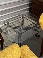 Front angled view of square glass coffee table with ornate black metal base and clear thick glass top.