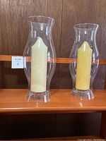 Two tall clear glass flutes each containing a pillar candle, displayed on shelf.