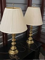 Pair of gold side table lamps with cream pleated shades on a black surface, showing bases and cords.