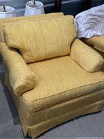 Front view of one yellow tweed upholstered living room chair showing cushioned seat, arms, back cushion, and skirted bottom edge on the floor.