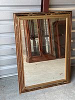 Full view of the rectangular mirror propped against a wall in a storage unit, showing its decorative frame with multiple rows of beaded inlays around the edges.