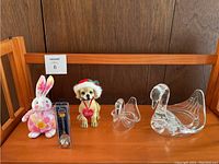 Five miscellaneous knickknack items on wooden shelf: stuffed bunny, dog figurine with Santa hat, packaged keychain, and two glass swans.