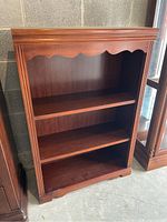 Front view of a dark brown wooden bookcase with three shelves and scalloped decorative design on the top front panel.