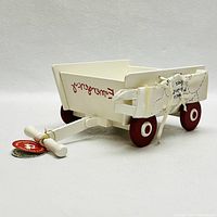 Side and front angled view of the cream colored wooden toy ambulance cart with red wheels, showing 'Emergency' script on the side and stretcher tied to the front with string.