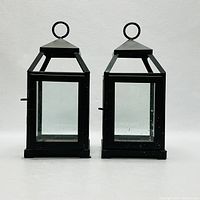 Front view of two rectangular black metal lanterns with glass panels and pyramid-shaped tops with round hanging rings.