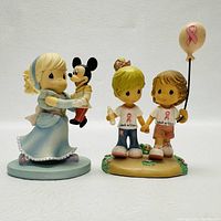 Front view of two Precious Moments collectible figurines: one with a girl holding a Mickey Mouse puppet, the other with two children holding hands and a pink ribbon balloon.