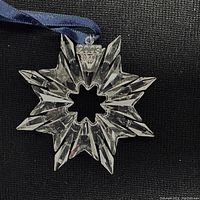 Crystal star ornament with 8 points, clear faceted crystal with metal top inscribed 2003 and attached blue ribbon on black background.