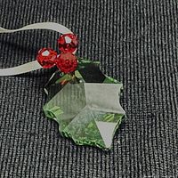 Close-up of green crystal holly leaf ornament with three red crystal berries and white ribbon on dark textured surface.