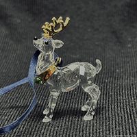 Side view of clear crystal reindeer figurine ornament showing faceted details, gold-tone antlers and green gem accents with attached blue hanging ribbon.