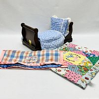 Front view of miniature wooden sleigh bed with blue floral quilted bedding and pillow, alongside two quilted blankets including a colorful patchwork and a pink-blue plaid blanket with Snoopy label.