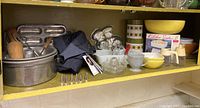 Wide shelf view showing an assortment of vintage baking items including pans, glass bowls, cookie cutters and a denim apron.