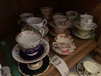 Photo showing multiple porcelain tea cups and saucers in assorted floral and decorative patterns on wooden shelf