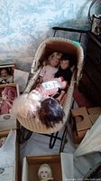 Photo showing a wicker doll pram with five vintage dolls inside and around it, some dolls seated inside the pram, others visible nearby in boxes, background includes floral wallpaper and room furniture.