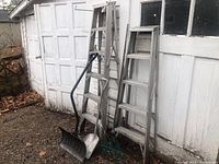 Two aluminum step ladders leaning against a white garage door with two shovels and one garden rake in front.
