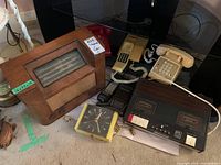 Full lot showing antique wooden radio with woven speaker, three vintage telephones (red rotary and beige/cream push-button), yellow wall clock, and black cassette answering machine on table.