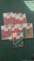 Eight Bardwil table linens in original packaging with poinsettia and Christmas bell designs arranged on a green carpet.