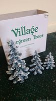 Three porcelain evergreen trees arranged outside the box showing snow-dusted branches, with the original packaging behind them.