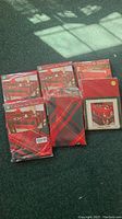 Photo showing seven new in package holiday table linens, mostly plaid in red and black with one solid red piece and one featuring holiday designs.
