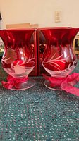 Photo of two ruby floral hurricane candle holders side by side with original boxes behind on carpeted floor.
