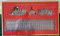 Close-up of pewter train cars and matching railroad tracks in original presentation box with red and gold background labeled Smithsonian Holiday Express Train.