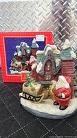 Photo of ceramic holiday figurine showing Santa Claus standing next to a snow-covered house with a sled full of toys and presents. The box with festive graphics is visible behind the figurine.