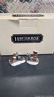 Photo showing both Christmas village figurines in front of original Hawthorne box on a black surface.