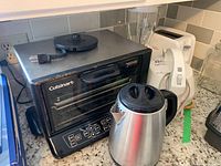 Cuisinart toaster oven, Black & Decker stainless steel kettle, and white toaster with Oster blender base seen behind them on kitchen counter.