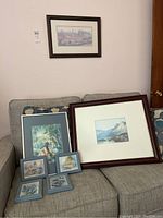View of seven framed prints on a couch including mountain landscape, woman under tree, and floral scenes.