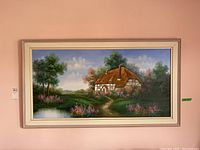 Framed and signed painting showing a thatched cottage with surrounding nature in a horizontal rectangular light frame, positioned against a beige wall.