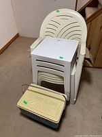 Photo of 4 stacked white plastic outdoor chairs with 3 stacked white plastic square tables on top, and a vintage Hot-N-Cooler portable cooler placed in front.