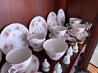 Photo showing fine bone china plates, cups, saucers, small cups, and a pitcher with pink floral patterns displayed on wooden shelves.