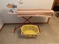 Photo shows striped ironing board with iron and box containing scissors and miscellaneous items on top. Two plastic laundry baskets underneath.