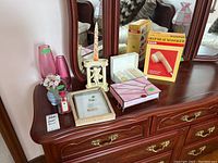 Wide view showing all items on a wooden dresser with mirror including two pink vases, white candlestick holder with twisted candle, stained glass box, small flower vase with pink flowers, framed artwork, small red bottle, and boxed massager.