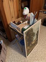 Photo of folding table holding various decor items including white ceramic dog figurine and small boxes, with framed landscape print leaning against it.