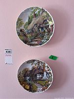 Front view of two decorative plates mounted on wall showing house and forest scenes in natural colors, each approx. 8 inches diameter