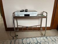 Full view of DVD player resting on metal frame stand with one shelf. Two remotes on top of the DVD player. Stand has caster wheels and metal tubular frame with brown shelf.