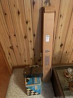 Tall cardboard box labeled as portable projector screen, next to smaller boxes and cords against wood-paneled wall.