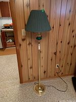 Full view of the brass floor lamp with green pleated shade positioned against wood paneling and tile floor in a basement room.