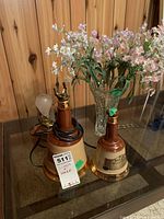 Photo showing two bell-shaped table lamps and a glass vase with artificial flowers on a wooden surface.