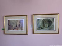 Two gold-framed prints hanging side by side on a pink wall showing interior architectural scenes with people.